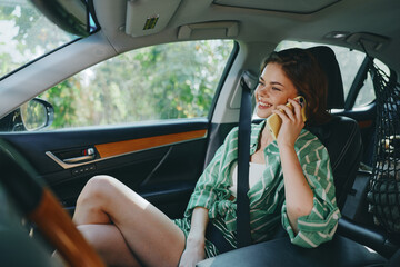 Young woman sitting in a car seat, smiling and talking on a smartphone, relaxed and stylish in...