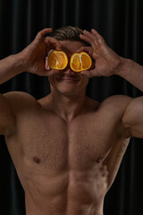 confident male figure with a muscular build holds two orange halves in front of his eyes smiling playfully. The dark background highlights his expression and physique.