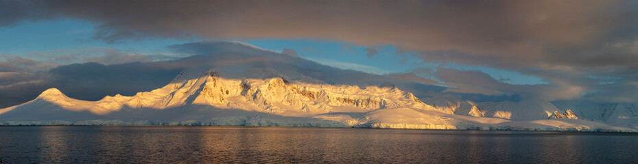Panoramic view of Antarctic mountains bathed in golden light during sunrise or sunset, with dark clouds and calm ocean waters creating a dramatic contrast. A breathtaking scene of the polar wilderness
