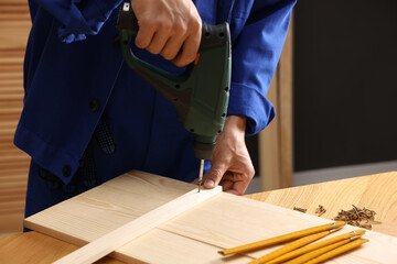 Professional repairman twisting screw into wooden plank with electric screwdriver indoors, closeup