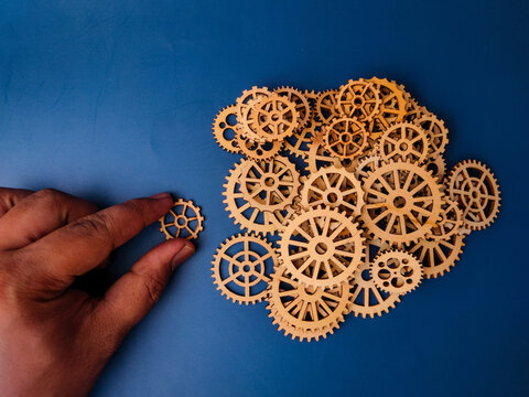 The image shows a hand placing a wooden gear, which is part of a larger arrangement of gears on a blue background
