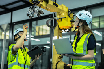 A team of two female engineers in hard hats works together on a large robot arm in a factory. They use a laptop and a controller to program the machine for its job. © ultramansk