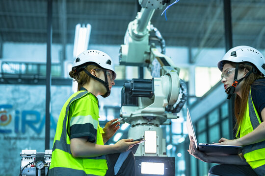 A female automation engineer calibrates a 6-axis industrial robotic arm on a production line. She uses a laptop to program its movement for an advanced manufacturing process.