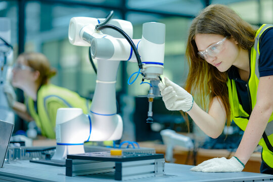 A team of two female engineers in hard hats works together on a large robot arm in a factory. They use a laptop and a controller to program the machine for its job.