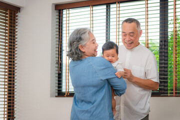 Fototapeta premium Asian grandparents holding and playing with their grandchild, Happy grandfather and grandmother laughing while holding a cute baby, Two generations of a family smiling and bonding together at home