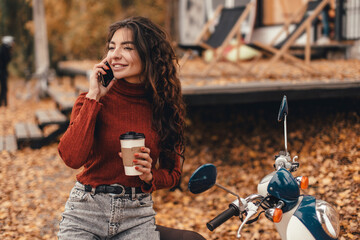 Beautiful woman in cozy outfit using smartphone or phone and drink coffee while sitting on chair near cafe in autumn park. Freelancer works remotely outside home.