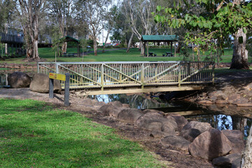 Fototapeta premium Bridge crossing over a narrow stream surrounded by large rocks, grass and trees at the Tyto Wetlands in Ingham, Queensland, Australia