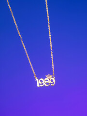 Close-up of a metallic gold pendant in the shape of "1989" on a chain, on a  purple background