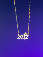 Close-up of a metallic gold pendant in the shape of "2012" on a chain, on a  purple background
