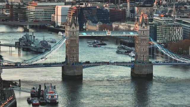 United Kingdom, London: Aerial view of Tower Bridge at sunset over River Thames with boats sailing and HMS Belfast docked, in London, United Kingdom. Drone flight footage