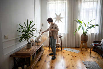 Calm tidy young woman using duster to collect dust from furniture while rearranging items at home. Home tasks, housewife duties, household chores in apartment, weekend responsibility, cleaning