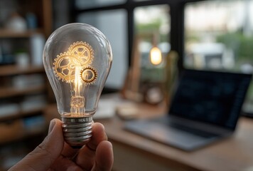 Lightbulb with gears inside held by a hand, laptop, shelf and bright window are blurred in background
