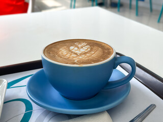 Close-up of a latte or cappuccino in a blue ceramic cup with symmetrical foam art, served on a tray in a modern café setting with stylish decor.