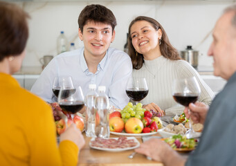 Friendly young girl and guy enjoying festive family dinner, spending time with elderly parents over wine and light snacks in cozy kitchen..