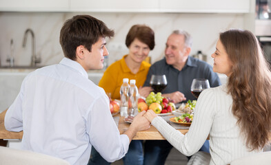 Couple sharing close moment at dining table, holding hands and locking eyes, while happy parents watching in background at family home. Blessing of parents
