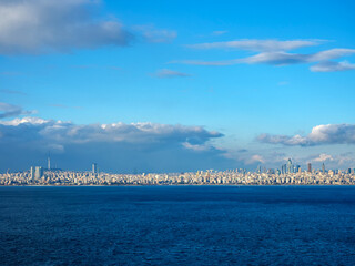 A distant city skyline is seen across a vast expanse of calm blue sea under a partly cloudy sky a panoramic landscape that blends urban life with the beauty of nature