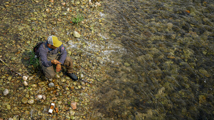 Fisherman wearing waders and baseball cap sitting on rocky river bank casting fishing line into clear water