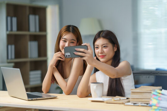 Two young businesswomen are taking a selfie with a smartphone in a modern office, smiling and enjoying a moment of fun during their workday