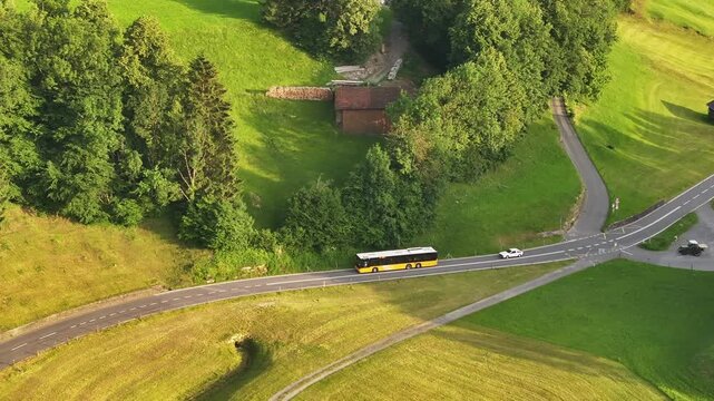 Aerial View Of Yellow Bus And Car Driving In The Road Along The Grassy Hill And Fields In Summer In Switzerland.