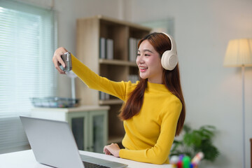 Smiling young woman taking selfie with smartphone and headphones while sitting at desk with laptop at home
