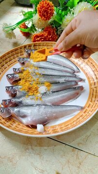 Vertical 1080x1920 stock video of seven fresh pabda (butter catfish) on a rattan-pattern plate. A spoon adds salt and turmeric for marination on a home kitchen counter&mdash;clean, appetizing food prep.