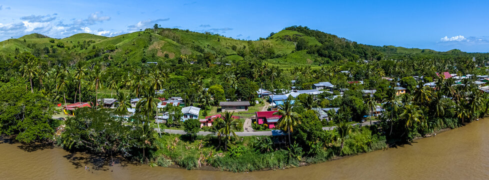 Panoramic aerial view of a traditional Fijian village with colorful houses nestled amongst palm trees on the banks of the muddy Sigatoka River, Fiji