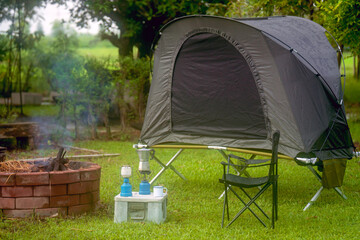 Camping tent and gas lantern on the grass next to a wood stove in the forest, with outdoor coffee-making facilities in a forest camping area