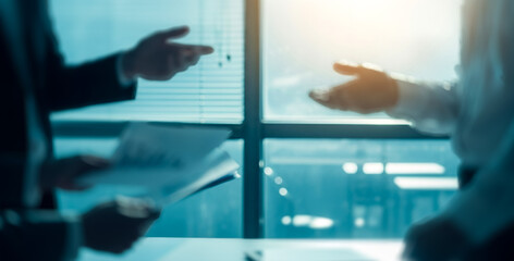 Two people in suits gesture during a business meeting near a window.
