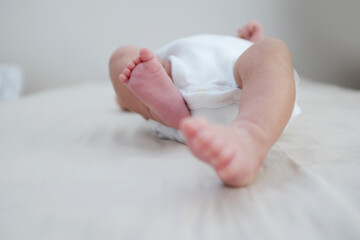 Gentle close-up of adorable newborn baby wearing white onesie, lying on soft bed with tiny feet raised in the air, minimal neutral background, tender lifestyle moment symbolizing innocence, family lov