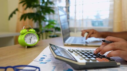 Office Desk with Calculator and Laptop, Person using a calculator and laptop at a desk with a green alarm clock - Powered by Adobe