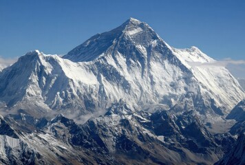 Fototapeta premium A majestic snow-covered mountain peak pierces the clear blue sky, surrounded by rugged terrain and lower, cloud-draped peaks in the distance