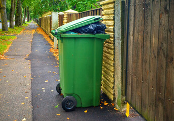 Green outdoor garbage bin with overflowing trash bag standing on a sidewalk near a wooden fence.