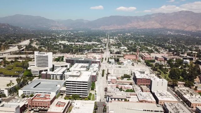 Drone pans above Pasadena, California, USA, showcasing the city skyline, busy freeway with fast moving traffic, modern buildings, and San Gabriel Mountains under a ​h​azy blue sky​ with fluffy clouds.