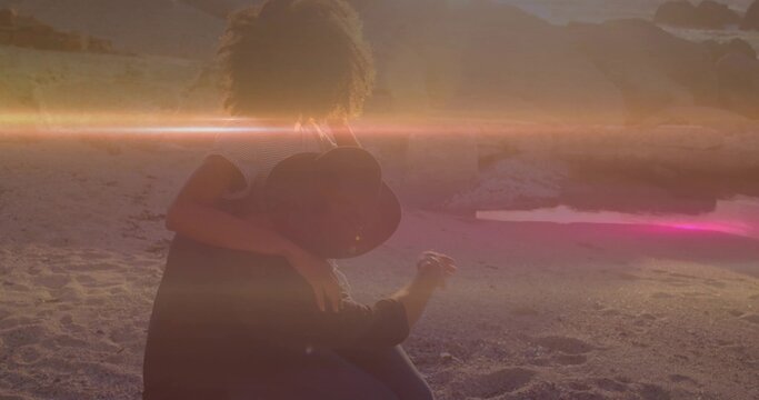 Hugging curly-haired woman and man wearing wide-brimmed hat sitting at water's edge at sunset