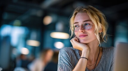 Fototapeta premium Young Caucasian woman with blonde hair and glasses sits at a desk, resting her chin on her hand, in a modern office environment with soft lighting.
