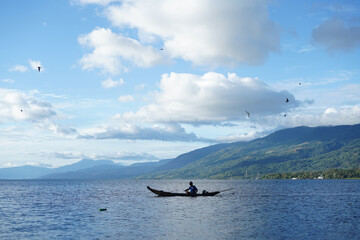Fototapeta premium Traditional fisherman on wooden canoe in calm lake with mountain background, blue sky, and flying birds, representing nature, lifestyle, environment, and peaceful rural travel scene.