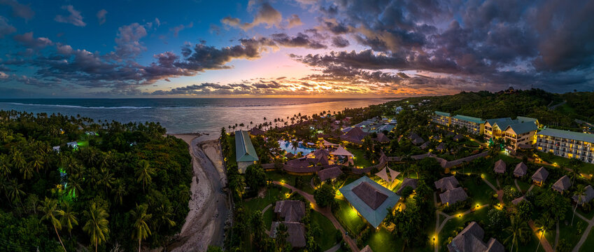 Aerial panorama of a luxury Fiji resort with lights on at twilight. The beautiful blue hour sky complements the illuminated pools, bures, and palm trees