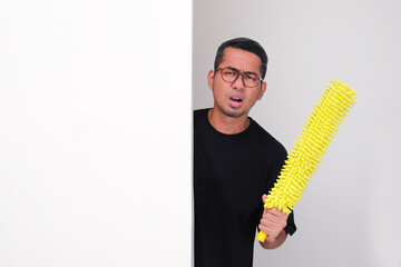 Adult asian man peeking behind wall holding duster with lazy face expression