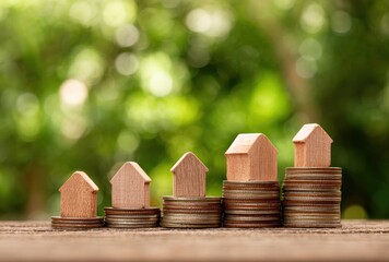 Wooden toy houses sit atop stacks of coins in a stair-step arrangement, blurry green bokeh background