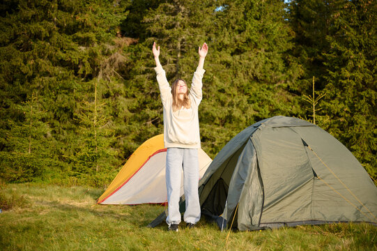 Woman hiker stretching at tent's entrance, warming up in the morning. Young female traveler at the tent looking out at a scenic view. Solo travel, adventure, and enjoying the outdoors.