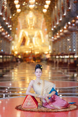 Fototapeta premium Elegant Thai pretty woman in traditional costume posing gracefully inside a golden temple hall.