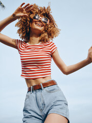 Joyful young woman dances outdoors in bright sunlight, wearing a striped crop top, denim shorts,...