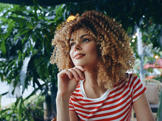 Portrait of a young woman with voluminous curly hair in a red and white striped tee, resting her chin on her hand amidst lush greenery and soft natural light.