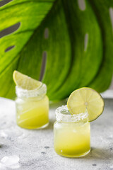 Two vibrant green shots of a cocktail, each garnished with a fresh slice of lime and a salt rim at monstera leaf Summer drink concept