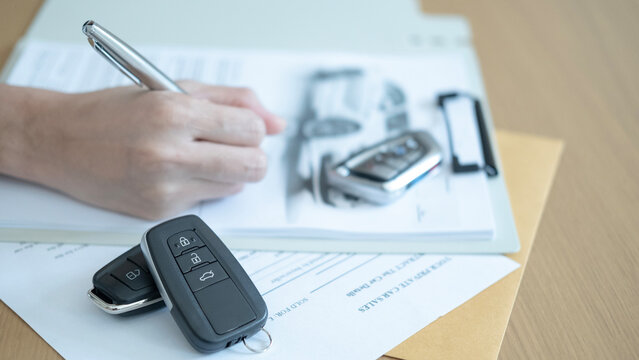 Hand signing car sales paperwork with keys on the table symbolizing agreement contract and transfer of vehicle ownership