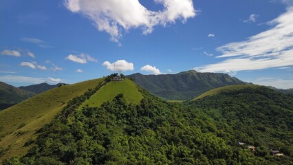 Wide aerial frame of rolling green hills around Mount Tapyas above Coron, Palawan, Philippines. A narrow ridge with antennas crowns the summit, with lush tropical forest under bright blue sky.