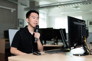 A young man sits in front of a desktop computer in a classroom computer lab, holding a microphone while speaking, responding thoughtfully during a discussion or presentation.