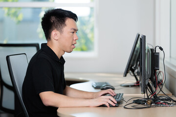 Fototapeta premium Young man types on a keyboard while focused on a desktop monitor in a computer lab classroom. Side profile view shows concentration in task.