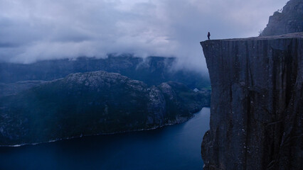 Captivating twilight view from Preikestolen Cliff, offering a dramatic perspective of Norways fjords and towering landscapes. A lone hiker stands on the precipice, embracing natures grandeur.