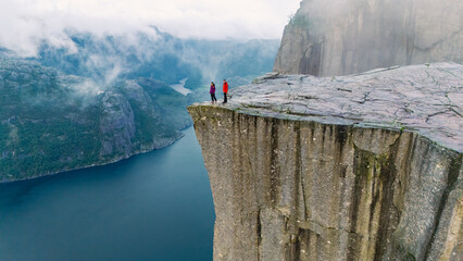 Two hikers stand on the edge of Preikestolen Cliff, overlooking a stunning fjord in Norway. The...